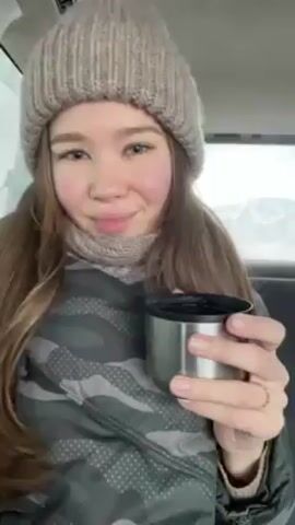 A young woman wearing a grey beanie, a scarf, and a camouflage jacket is sitting in the back of a car. She is holding a silver mug, and the camera pans to show a small dog wearing a black harness sitting on the seat next to her. The dog looks at the camera. The car is in motion, and snow-covered mountains are visible outside the window. Another person, who is blonde, is sitting in the front passenger seat.