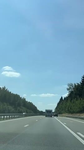 The video shows a first-person view of driving on a highway on a clear day. The sky is blue with a few scattered clouds. Trees line both sides of the highway, with a metal guardrail on the left. The highway has multiple lanes and white lane markings. In the distance, a white recreational vehicle (RV) is driving in the same direction. The RV appears to be a camper van or a small motorhome. The overall environment is a rural or semi-rural setting.