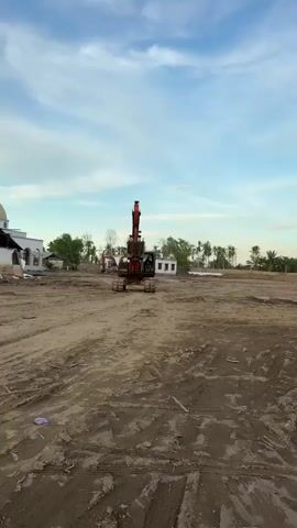 An excavator with a driver is shown in a construction site. The excavator is red and black and has tracks. The driver is wearing a dark shirt and is sitting in the cab of the excavator. The construction site is a dirt field with some trees and buildings in the background. The sky is blue with some clouds.