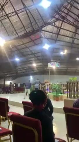 The video shows the inside of a large building, seemingly a church, with a high, metal ceiling. The room is illuminated by bright, overhead lights and a purplish light towards the stage. The focus is on the front of the room, where a raised stage area is adorned with decorative plants and a lectern where a person in light clothing speaks. Several people are standing or walking around on the stage. The foreground shows rows of red chairs filled with people who are watching the events unfold on the stage. The atmosphere seems calm and focused on the activity at the front.