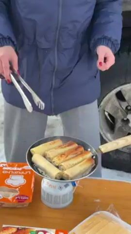 A person wearing a blue jacket and grey pants is cooking rolled crepes in a frying pan with wooden handle. They are standing outdoors on a snowy ground near a car. A small portable propane stove is underneath the frying pan on a wooden table. The person is using metal tongs to turn the crepes in the pan. There are two packages of crepes on the table.
