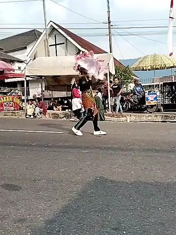 A group of people in costumes are parading down a street. There are buildings and tents along the side of the street, and a crowd of onlookers. The person in the foreground is wearing a black and brown outfit with green stripes down the legs, a headdress, and white shoes. They are holding a pink and white feathery object. To their left, another person in a red top and white skirt is holding a similar feathery object. Further back, more people in costumes can be seen. The street is paved, and there are power lines overhead.