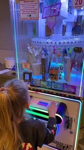 A young girl with blonde hair in a ponytail stands in front of a brightly lit arcade claw machine, viewed from behind. She places her hand on the machine's joystick, appearing focused on controlling the claw. The machine is filled with various prizes, including small boxes, erasers, and what appears to be a digital watch. A sign on the machine details the rules of play. The environment suggests an indoor entertainment area.