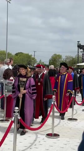 The video shows a line of academics in full regalia walking along a white tiled path guided by red rope barriers. The academics wear a variety of gowns in colors like purple, red, black, and blue, each decorated with stripes or sashes. They also wear caps, some with tassels and others with flat tops. The path is lined with people, many of whom are looking at the academics. In the background, there is a cloudy sky, trees, power lines, and a stage with a large screen.