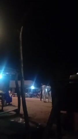 The video shows an outdoor scene at night, illuminated by artificial lights. In the foreground, a rudimentary wooden shelter with a roof is visible, with tables and chairs underneath. A person in a blue garment is seated at one of the tables. To the right of the shelter, a tall wooden pole stands vertically. Further back, a white truck with a large red tarpaulin covering its cargo area is parked. Several people, appearing to be male and wearing light-colored clothing, are standing near the truck. The ground appears to be dirt or gravel. The overall ambiance suggests a roadside stop or a gathering place, possibly a market or rest stop at night. The lighting casts strong shadows, and some lights create lens flare effects.