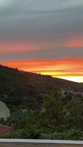 A time-lapse video shows a vibrant sunset over a hillside town. The sky transitions from a soft gray to streaks of orange, yellow, and red. Below the sky, a green, tree-covered hill slopes down to a cluster of houses with red-tiled roofs. The houses are nestled together, with some lights beginning to appear. The foreground features lush green foliage and a white railing.
