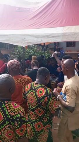 A group of people are gathered under a white and red tent. Most of the people are facing the same direction, with several wearing patterned shirts. Some are standing, some are talking to each other, and one person is holding a white bottle. The environment appears to be outdoors in a residential area.