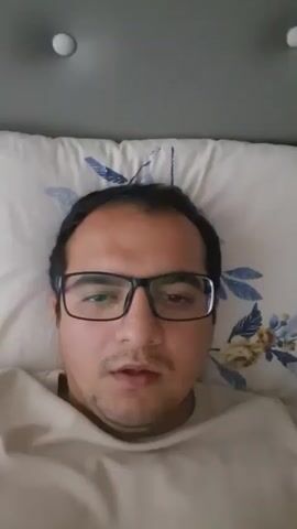 A close-up, high-angle, point-of-view shot shows a man with dark hair, glasses, and a light-colored shirt lying on a pillow with a floral pattern. The man's eyes are closed at the beginning of the video. He then opens his eyes and smiles. The background shows a grey headboard with two white buttons. The video is brief and appears to be a personal recording.