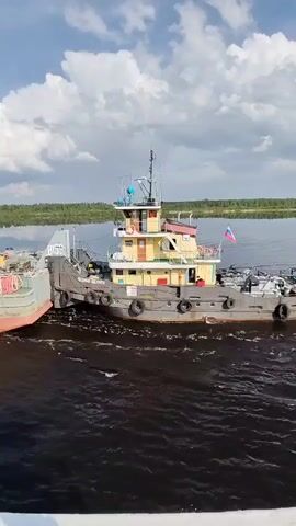 The video shows a tugboat moving through a waterway on a partly cloudy day. The tugboat is painted in a combination of yellow, gray, and red colors, and it is towing a barge alongside it. The Russian flag is visible on the tugboat. The water is dark and reflects the sky. The background shows a green shoreline with trees.