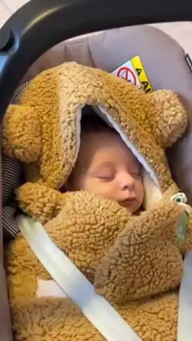 The video shows a baby sleeping in a car seat. The baby is wrapped in a brown, hooded, fuzzy outfit that resembles a teddy bear. The baby is secured in the car seat with a white harness. The background is out of focus, but the headrest of the car seat and a warning label are visible.  The camera zooms slightly in on the baby's face. There are indistinct voices in the background.
