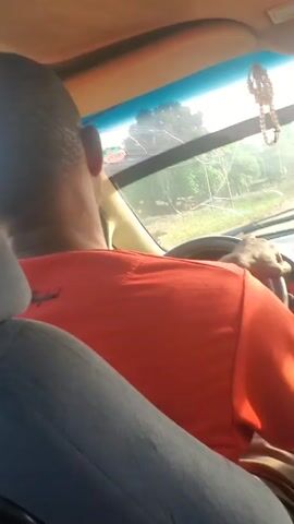 A man in an orange t-shirt is driving a car. The camera appears to be positioned behind the driver, focusing on the back of his head and shoulders. Outside the windshield, there is a view of a rural road with some structures and vegetation visible. The car's interior includes a gray seat.
