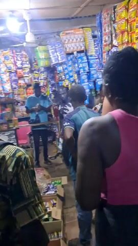 The video shows the interior of a small store, tightly packed with merchandise displayed on shelves. Several individuals are present, including a woman in a pink tank top and dark pants standing with her hands on her hips, seemingly observing the scene. Other customers are browsing or standing near the counter. The store appears to be selling various snacks, drinks, and household items. The lighting is provided by fluorescent fixtures, giving the store a brightly lit appearance.