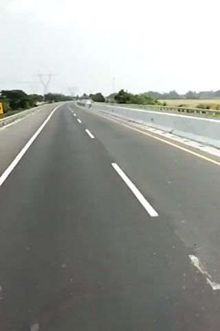The video shows a POV shot of a car traveling on a highway. The highway is paved with asphalt and has white lane markings. There are guardrails on both sides of the road, and trees and power lines can be seen in the background. The sky is overcast. A few other cars can be seen in the distance.