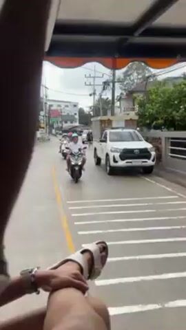 The video shows a person riding in the back of a vehicle with an open-air top, likely a tuk-tuk or similar transportation. The person's legs, feet in white sandals, and lower arm with a watch are visible in the lower portion of the frame. The camera faces forward, capturing the street ahead. The street has white parking stripes on one side and features a row of buildings on both sides. The sky is overcast.

Several people on motorbikes are driving on the road in the same direction. A white Toyota pickup truck is visible, parked along the side of the road. Utility poles with electrical wires run along the side of the street. There is moderate traffic. Other vehicles can be seen further down the road. The overall impression is a street scene in a tropical or Southeast Asian location. The person in the vehicle occasionally grabs the handrail for support.