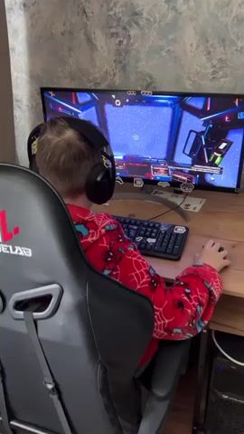 Here's a description of the video:

A young boy is playing a video game on a computer. He is wearing headphones and a red Spiderman onesie, seated in a black gaming chair. The game displays a space scene with a blue background and stars. 

The desk is wooden and cluttered with stickers, including a "Metallica" logo and a "low battery coffee cup". A black keyboard and a mouse are visible, being used by the boy. There's also a decorative metal arc propped behind the keyboard. A coin is visible on the desk; it features the words "Санкт-Петербург" (Saint Petersburg) around the top and "Победа в Мире" (Victory in the World) around the bottom. The wall behind the monitor has a gray textured pattern.