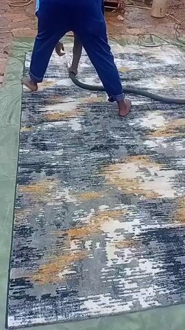 A person wearing blue pants is cleaning a rug with a hose and a machine. The rug has a gray, black, white, and yellow abstract pattern. The person is standing on the rug, holding the hose, and directing it across the surface. The machine, which appears to be a carpet cleaner, is to the right of the rug and connected to the hose. The rug is laid out on a green tarp on a paved surface.