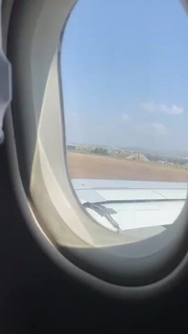 The video shows a view from an airplane window. The window frame is visible in the foreground, with the interior of the aircraft on the left side. Outside the window, an airplane wing is visible, taking up the lower half of the frame. Beyond the wing, a landscape is visible, consisting of a flat, brown expanse with some lighter colored areas. In the distance, there are some low hills and buildings. The sky is clear and blue with a few scattered white clouds. The plane appears to be on the ground, possibly at an airport or preparing for takeoff or landing. The lighting suggests it is daytime. There are no people or specific actions clearly discernible in the video.