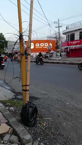 The video shows a street scene with a medium level of traffic consisting primarily of motorbikes. An orange sign is visible on a building across the street, reading "GODA, SAFER SMART TV." In the foreground, there is a bamboo structure wrapped with wires, suggesting it might be a makeshift utility pole. At the base of this structure is a black garbage bag. A car with green rims also drives by.
