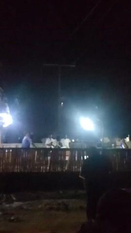 The video is taken at night and shows a gathering of people behind a low fence or railing, illuminated by bright lights. The darkness makes it difficult to distinguish individual features, but the general shapes of people are visible. Above the crowd, power lines stretch across the sky. The ground in the foreground appears uneven and somewhat rough. The overall atmosphere seems lively, possibly suggesting an event or social gathering.