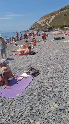 This is a video of a crowded pebble beach on a sunny day. There are many people sunbathing on towels and in the water. Some people are swimming, others are wading, and some are playing. There are also a few umbrellas providing shade. In the background, there are boats and a steep, rocky hill. The water is a clear blue. The overall atmosphere is one of leisure and recreation.