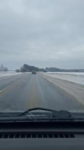 A driver's-eye view of a two-lane highway with one lane in each direction. The road is black and wet, with some bare spots.  Snow is piled on the side of the road. There is a line of trees on the left and a field of snow on the right. The sky is overcast.  A white car passes in the opposite direction.  There are two cars driving ahead in the same direction.