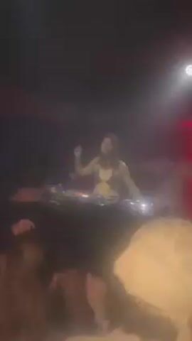 The video shows a dark nightclub environment. A female DJ, dressed in a black top, is visible behind a DJ console, performing and dancing. Several people are gathered in front of the console, watching her. The lighting is dark, with a spotlight and red lights illuminating the DJ and the surrounding area. Some of the crowd members are raising their hands and appear to be dancing.