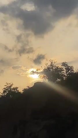 The video captures a serene natural landscape at either sunrise or sunset in Ufueseni, Nigeria. The scene is dominated by a bright sun, partially obscured by clouds, casting radiant golden rays across the sky and through the silhouetted trees and hills in the foreground. The sky transitions from a warm, bright yellow and orange hue near the horizon, where the sun is located, to lighter and darker grey clouds above. The lower portion of the frame shows the dark, silhouetted outlines of hills or elevated terrain covered with sparse trees and foliage, appearing almost black against the luminous sky. No people or artificial objects are visible. The atmosphere is calm and contemplative, highlighting the beauty of natural light and silhouettes.