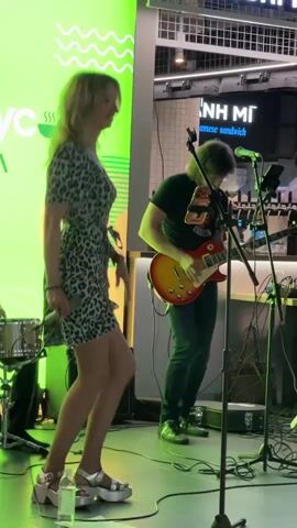 A young woman with blonde hair, wearing a short leopard-print dress and silver platform sandals, stands center-left, holding a microphone in her right hand and singing. She is performing on a stage with a green backdrop featuring white wavy lines and a logo. To her right, a person with shoulder-length brown hair, wearing a black t-shirt and dark jeans, plays a red and yellow electric guitar. They are both illuminated by stage lighting. In the background, there is a bar area with bottles and patrons, and a sign that reads "Banh Mi Japanese Sandwich." Other musical equipment, including a drum set and amplifiers, are visible on the stage. The overall environment appears to be an indoor venue, possibly a bar or restaurant.