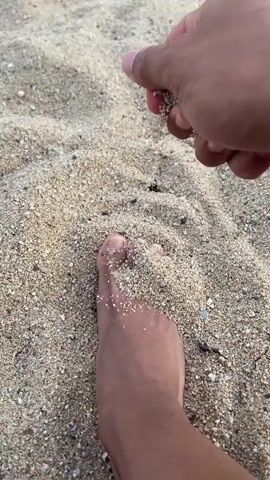 The video shows a close-up of a person's foot partially submerged in sand on a beach. A hand is seen above the foot, letting sand fall onto the foot and surrounding area. The sand is a light beige color with small pebbles and shells mixed in. The foot is bare and appears to be light-skinned. The overall lighting suggests an outdoor, daytime setting.