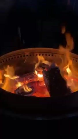 The video shows a fire burning inside a black metal fire pit. The fire pit has a series of small holes near the top edge. The fire is composed of several pieces of wood. The wood is charred and glowing red in the center. Flames are flickering above the wood. The surroundings are dark, suggesting the video was taken at night or in a dimly lit place. In the background, a person speaks.