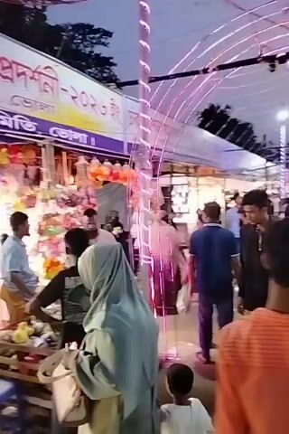 The video captures a lively night market or fair scene in Charfasson, Bangladesh. Numerous people, including men, women, and children, are seen walking, browsing, and shopping among brightly lit stalls. The stalls display various goods, appearing to include bags, accessories, and other small items. The area is illuminated by many string lights and decorative arches, creating a festive atmosphere. The time of day is clearly night, with artificial lighting prominently featured. A banner above the stalls displays Bengali text, which includes phrases like "বাণিজ্য মেলা" (Trade Fair), "ভোলা" (Bhola), "আয়োজনে: চরফ্যাশন" (Organized by: Charfasson), and "বাস মালিক" (Bus Owners).