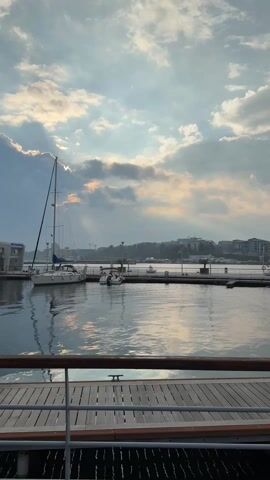 The camera, positioned behind a wooden railing, captures a tranquil waterfront scene. A sailboat is docked, its mast reaching toward a sky filled with scattered clouds and sunbeams. The water reflects the sky's light, creating a shimmering effect. A pier extends into the water, lined with street lamps. In the distance, buildings and construction cranes are visible on the shore. The atmosphere is calm and peaceful.