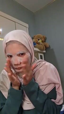 A woman wearing a hijab and a sheet mask on her face is the primary focus of the video. She adjusts the mask, smoothing it onto her face with her hands. The woman then holds up a pink box with the words "SKINTIFIC NIACINAMIDE Brightening Daily Mask" printed on it, looking directly at the camera. The background features a white cabinet, a light gray wall, and a shelf with a teddy bear on it. She is wearing a green and pink dress. The lighting is somewhat soft, creating a muted color palette.