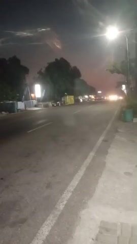 The video shows a nighttime view of a street from the perspective of someone walking along the sidewalk. A streetlamp illuminates the scene, casting a bright light on the asphalt road and the sidewalk. The road is mostly empty, with a few cars in the distance, their headlights creating streaks of light. Trees line the side of the road, their leaves dark against the night sky. There are buildings and signs visible on the left side of the road, but they are blurry and indistinct due to the low light conditions and the movement of the camera. The sky is dark with some clouds, and there appears to be some light pollution or a haze in the distance. The overall atmosphere is calm and quiet, with the sounds of distant traffic being the only discernible noise. The video is shaky, suggesting it was recorded handheld.