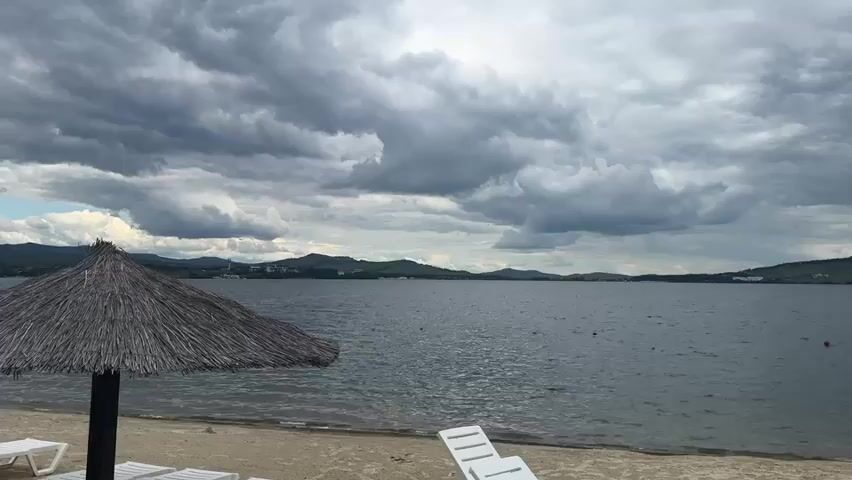 The video shows a beach scene on a cloudy day. In the foreground, there are two white lounge chairs and a thatched umbrella providing shade on the sand. The beach meets a large body of water with gentle waves. In the distance, there are rolling hills and mountains under a dramatic, cloudy sky. The water has several buoys or markers scattered across it. On the right side of the video, a small wooden dock is visible with a few people and a boat. The overall impression is a peaceful, somewhat overcast day at a lake or seaside resort.