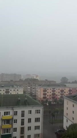 The video shows an aerial view of a city on a foggy day. The buildings are mostly apartment complexes with colorful facades. There are trees lining the streets and a few cars visible. The weather appears to be overcast and possibly rainy, as the streets look wet. The fog obscures the distance, making it difficult to see beyond the immediate cityscape.