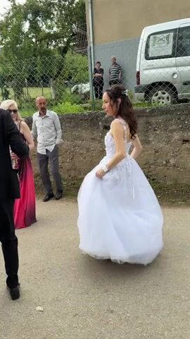 The video shows a bride in a white gown dancing with two other people on a paved path. The bride has long, curly brown hair, and her dress has floral embellishments. A man in a black suit and a woman in a maroon dress are holding her hands and dancing with her. The woman in the maroon dress is wearing a pink shawl. Another woman in a pink dress and sunglasses stands nearby, looking on. In the background, two men are standing near a chain-link fence and a white van is parked. The setting appears to be a residential area with a concrete wall and greenery visible. The atmosphere seems celebratory and joyful as the bride twirls in her dress.