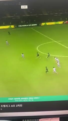 A video capture of a soccer game shows several players on a green field. The game appears to be in progress, with players moving the ball. The background displays advertisements and text indicating it is a FIFA World Cup 2026 event. The lower part of the screen shows additional text and branding.