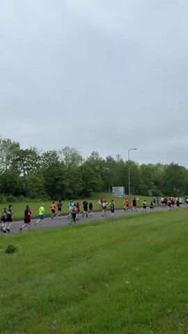 The video shows a group of people running a race on a paved road. The runners are of various ages and wear different colored athletic clothing. On the right side of the road is a grassy field, and on the left is a row of trees. A road sign is visible in the distance. The sky is overcast. The runners are moving from left to right across the screen.