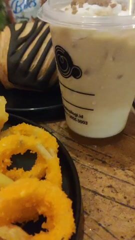 The video displays a close-up shot of a table laden with food and drinks. In the foreground, a black plate holds a generous portion of french fries, sprinkled with herbs. Next to the fries are several golden-brown, ring-shaped onion rings, also dusted with seasoning. Further to the left, a scattering of fried wontons and some sliced sausages add to the savory selection. A small dollop of white sauce and a pool of red sauce are visible at the bottom of the plate. In the background, a croissant with dark chocolate drizzle sits on another black plate. To the right, a clear plastic cup with a white lid contains a creamy, light-colored beverage, possibly coffee or a milkshake, topped with chocolate shavings. Another green drink in a similar cup is partially visible to the far left, and a grey plate with white, puffy snacks is in the mid-ground. The lighting suggests an indoor setting, likely a restaurant or cafe. The camera angle is low and slightly tilted, focusing on the food items.