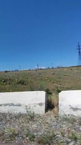 The video displays an outdoor, sunny scene under a clear blue sky. The camera moves horizontally across a grassy, uneven terrain, revealing dry, brown vegetation mixed with green patches and scattered rocks. In the distance, several utility poles are visible against the horizon. A large, white, rectangular object briefly appears in the foreground on the left before moving out of frame, suggesting motion from a vehicle or similar moving platform.