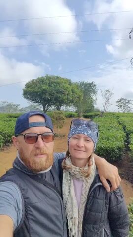 A selfie-style video shows a man and a woman outdoors, standing in what appears to be a tea plantation. The man, on the left, has a beard and is wearing a dark blue baseball cap, sunglasses, and a vest. The woman, on the right, is wearing a patterned headscarf, sunglasses, and a black jacket. The man has his arm around the woman. In the background, there are rows of tea bushes and a dirt path. Further back, there are trees and mountains visible under a partly cloudy sky. The man speaks at the beginning of the video, then he and the woman pose for the camera, the woman making a peace sign.