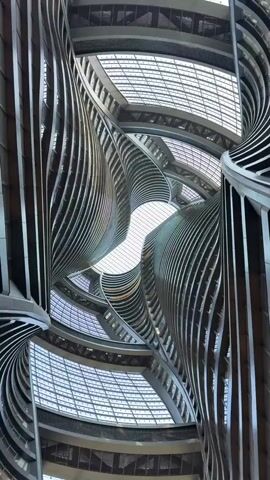 The video shows an interior view looking up at the architecture of a building. The architecture consists of curved, layered levels with geometric patterns, creating a spiral effect. The ceiling appears to be glass or transparent, allowing light to enter. A person wearing a dark cap appears briefly in the foreground.