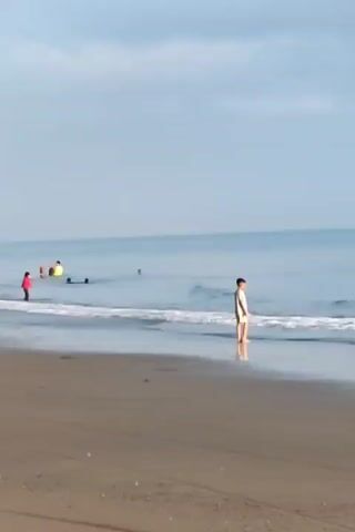 The scene is a beach on a cloudy day. The sand is dark. There are several people in the water; some are swimming and some are standing in the shallow water. A person in a white shirt and shorts stands at the edge of the water near the right of the frame. Waves gently break along the shoreline. The ocean is calm. The sky is overcast, but the horizon is visible.
