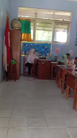 The video shows a classroom with students sitting at desks. A teacher, wearing a white hijab and black pants, stands at the front of the classroom. Some students are wearing green uniforms and hijabs. There are windows with green and yellow curtains, a world globe, a clock on the wall, and various decorations on the walls. The floor is tiled. The students appear to be listening to the teacher.
