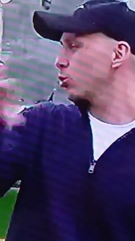 The video begins with a close-up of a person wearing a dark baseball cap and a purple zippered jacket over a white t-shirt. The person appears to be speaking, with their mouth open. In the blurred background, another person is visible, wearing a dark t-shirt and looking away from the camera. The setting appears to be outdoors, possibly near a stage or event setup. The video then cuts to a close-up of a white dog's eye and snout, showing pink gums and teeth. The dog appears to be panting or possibly injured, with a reddish substance visible on its muzzle. The background is dark and indistinct. The overall impression is a transition from a human subject to an animal subject.