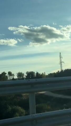 The video pans across a landscape with a blue sky dotted with white clouds. In the foreground, a silver guardrail runs horizontally across the bottom of the frame. Beyond the guardrail, there is a grassy area and then a dense line of green trees. In the distance, several structures are visible including what appears to be a tall, lattice-like tower, possibly a transmission tower or a derrick. There are also glimpses of more trees and rolling hills. The overall impression is a rural or roadside scene on a clear day.