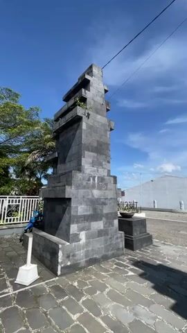 The video shows an architectural stone monument in the foreground. It's comprised of stacked stone blocks with a rough, dark grey finish. The camera moves to the right, revealing a paved plaza. In the distance, a prominent mountain is partially shrouded in clouds against a blue sky. A white building and a communication tower are also visible. Two people on a scooter drive from left to right across the plaza.