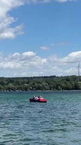 A red paddle boat with at least three people on it is moving across a body of water. The water is a greenish-blue and has small waves. In the background, there is a line of trees on the shore, and a tall wind turbine is visible above the trees on the right side of the frame. The sky is blue with scattered white clouds.