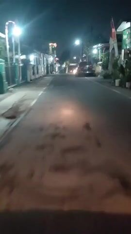 The video shows a low-angle perspective of a street at night. The camera is moving, creating a blurred effect. On the left side of the road, there are buildings with lights and flags displayed. On the right side of the road, there are also buildings and a car is visible. Streetlights illuminate the road. The overall impression is that of a residential or commercial street in a town or village.